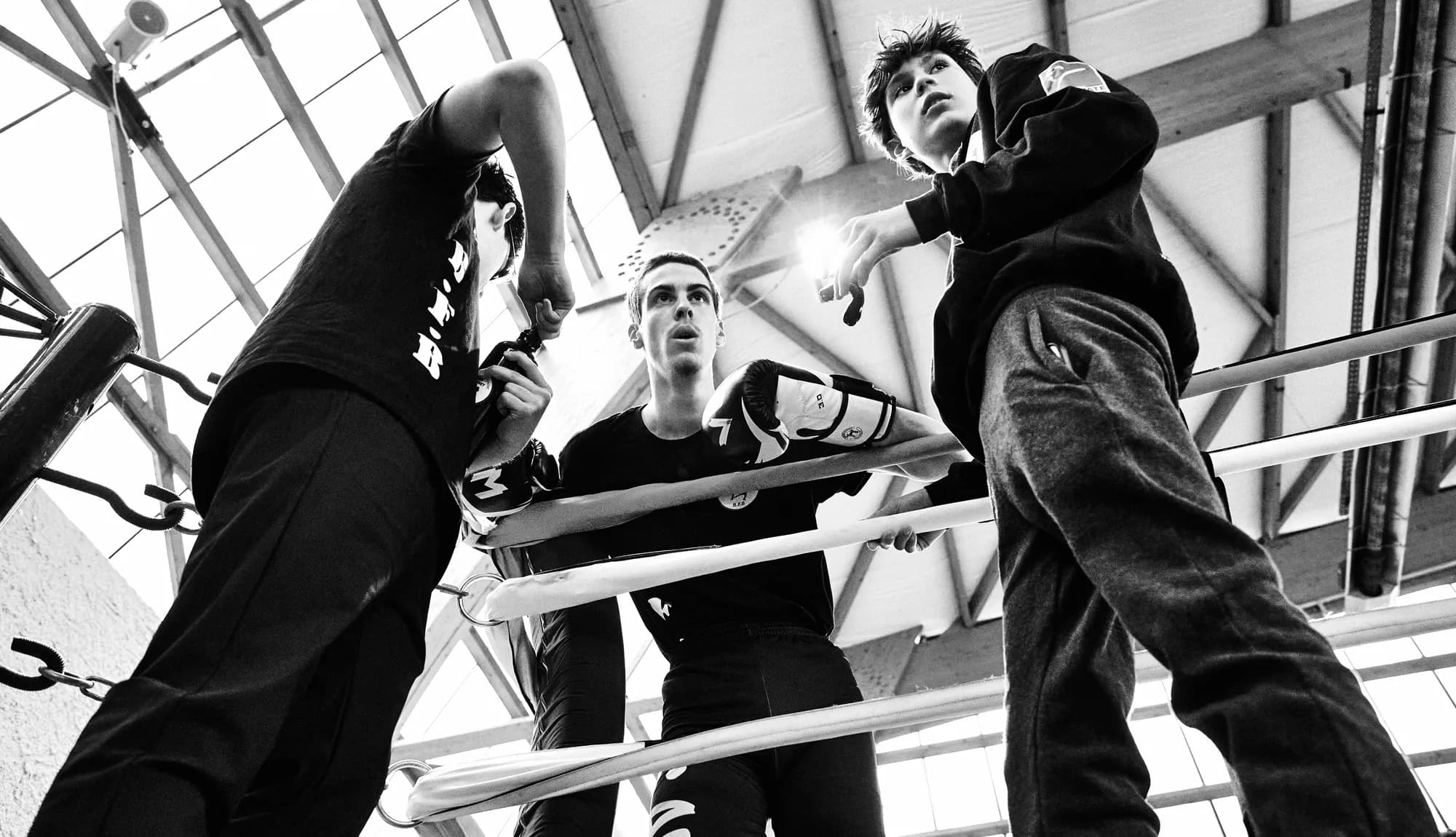 Deux entraîneurs et un jeune boxeur accoudés aux cordes du ring, vus en contre-plongée